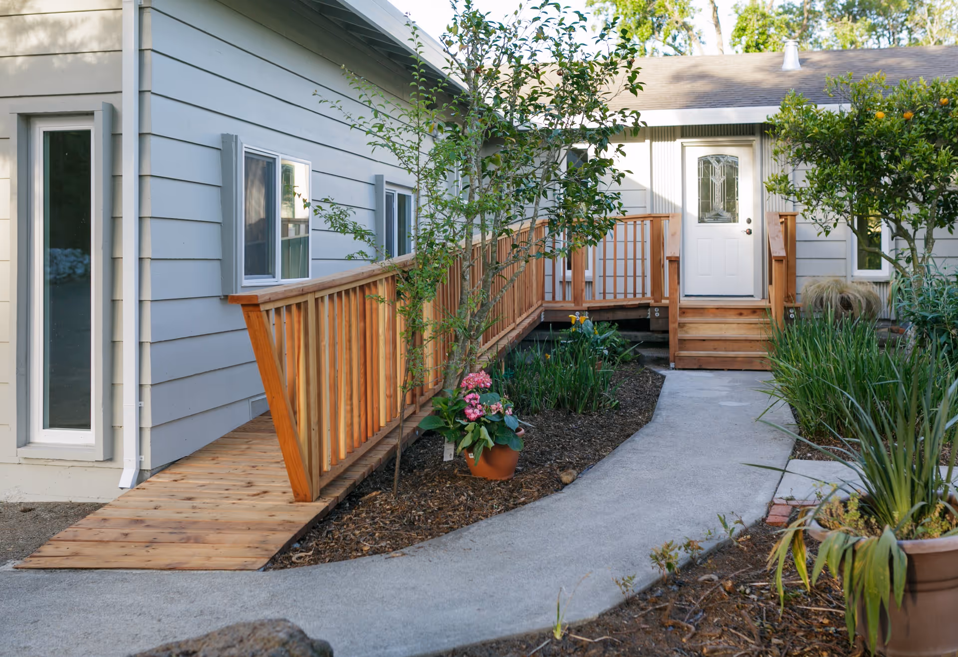 Outdoor view of a senior living facility entrance with a wooden ramp and stairs leading to a white door. The area is landscaped with plants, flowers, and trees along a curved concrete pathway.