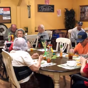 Several elderly residents seated around communal dining tables eating and socializing in a dining room.