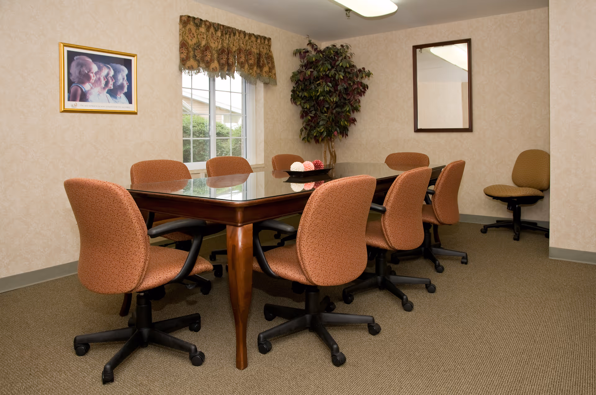 Meeting room with a glass-topped wooden table surrounded by upholstered swivel chairs, a window, wall art, and a potted plant.