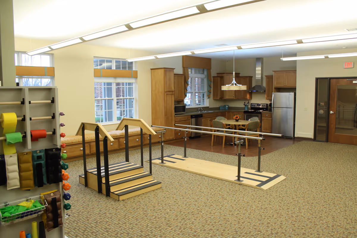 Spacious rehabilitation therapy room with parallel bars and practice stairs in front of a kitchenette and seating area.