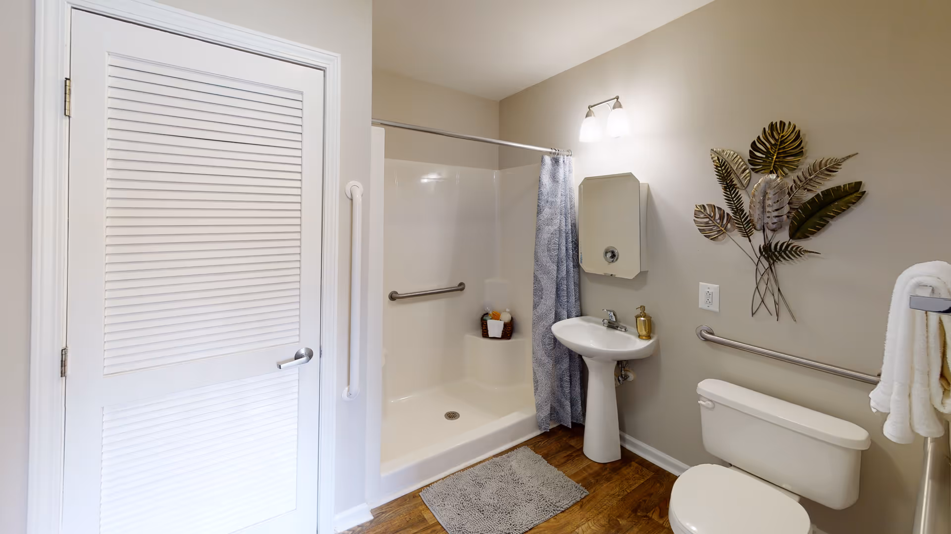 Accessible bathroom with a walk-in shower, pedestal sink, toilet, grab bars, and decorative wall art on wood-look flooring.