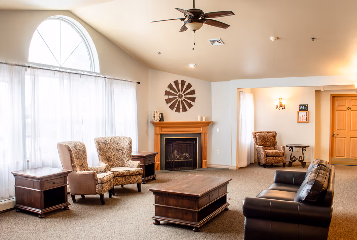 A cozy senior living facility common area with a large arched window covered by sheer white curtains, two patterned armchairs, a wooden coffee table, a black leather sofa, a wooden fireplace with decorative wall art above it, and a ceiling fan. There is also a small side table and another armchair near a doorway with a wooden door numbered 24.