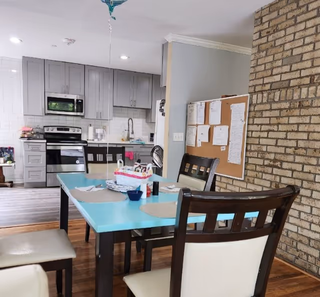 Interior view of a dining area with a turquoise table set with placemats, napkins, and condiments. Four chairs with dark wooden frames and light cushions surround the table. In the background, there is a modern kitchen with gray cabinets, a stainless steel oven, microwave, and a sink. A bulletin board with papers is mounted on a wall next to a brick accent wall.