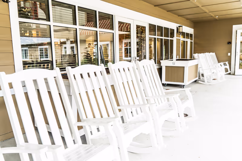 A row of white wooden rocking chairs lined up on a covered porch outside a building with large windows reflecting the surroundings.