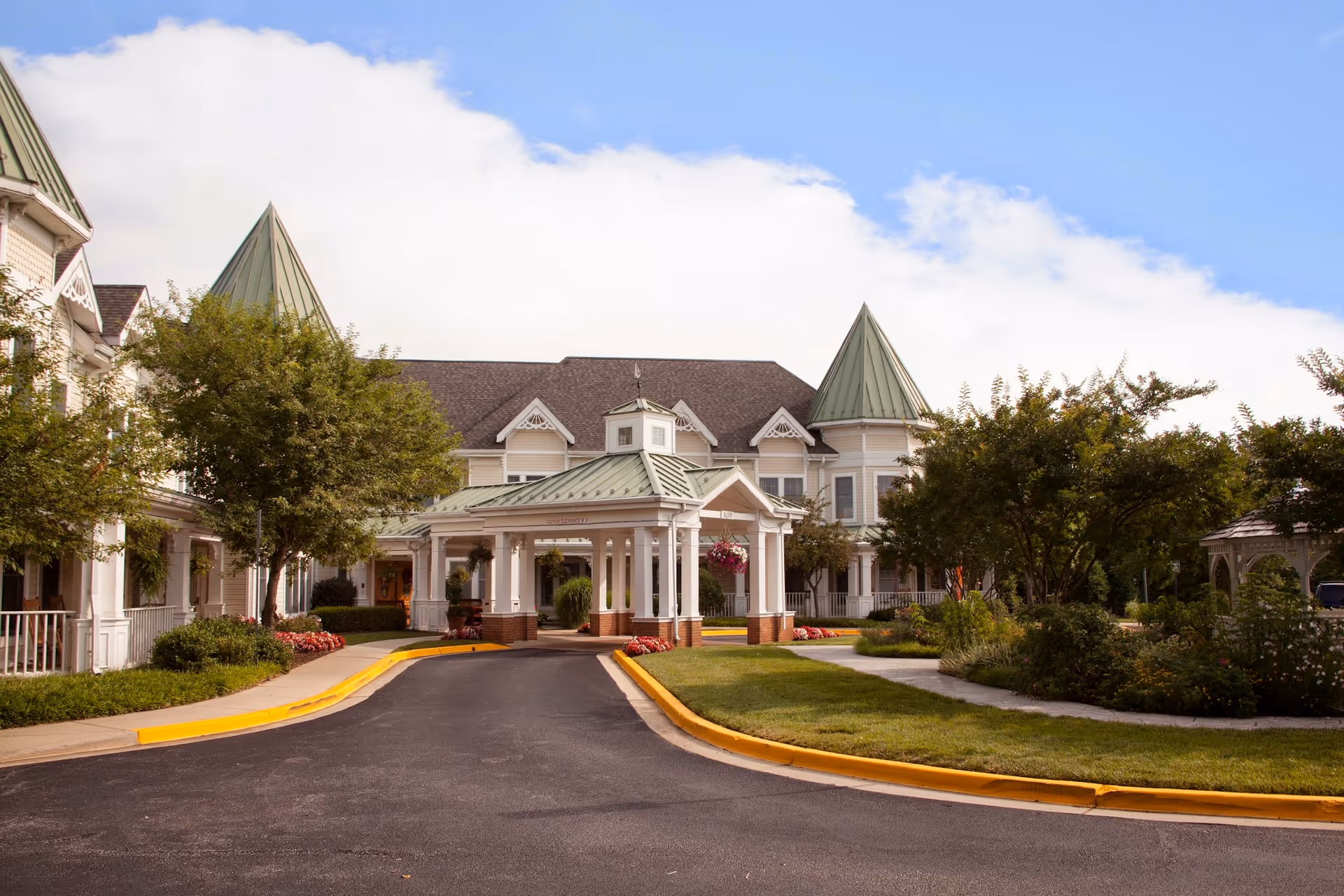 Front exterior view of a senior living facility with a covered entrance, manicured landscaping, trees, and a clear blue sky with some clouds.