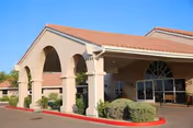 Exterior view of a building with a covered entrance supported by large columns, beige walls, and a red-tiled roof under a clear blue sky. There are bushes and a red curb in front of the entrance.