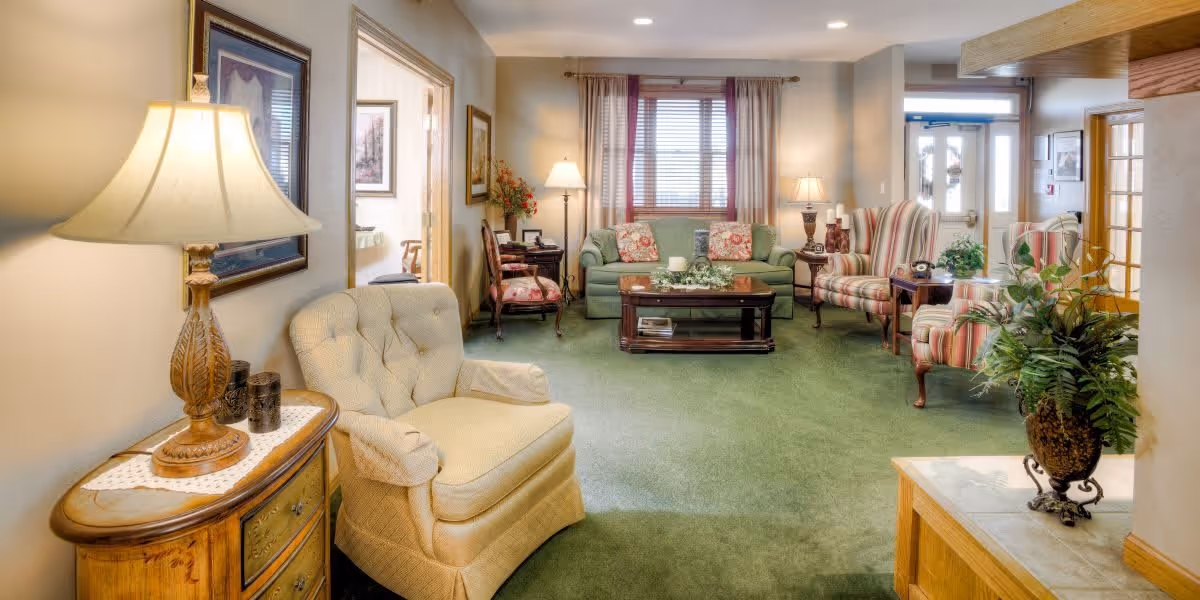 A cozy living room with green carpet, a beige armchair, a wooden side table with a lamp, a green sofa with floral cushions, two striped armchairs, a wooden coffee table with decorative items, and several lamps providing warm lighting. There are framed pictures on the walls and a window with curtains letting in natural light.