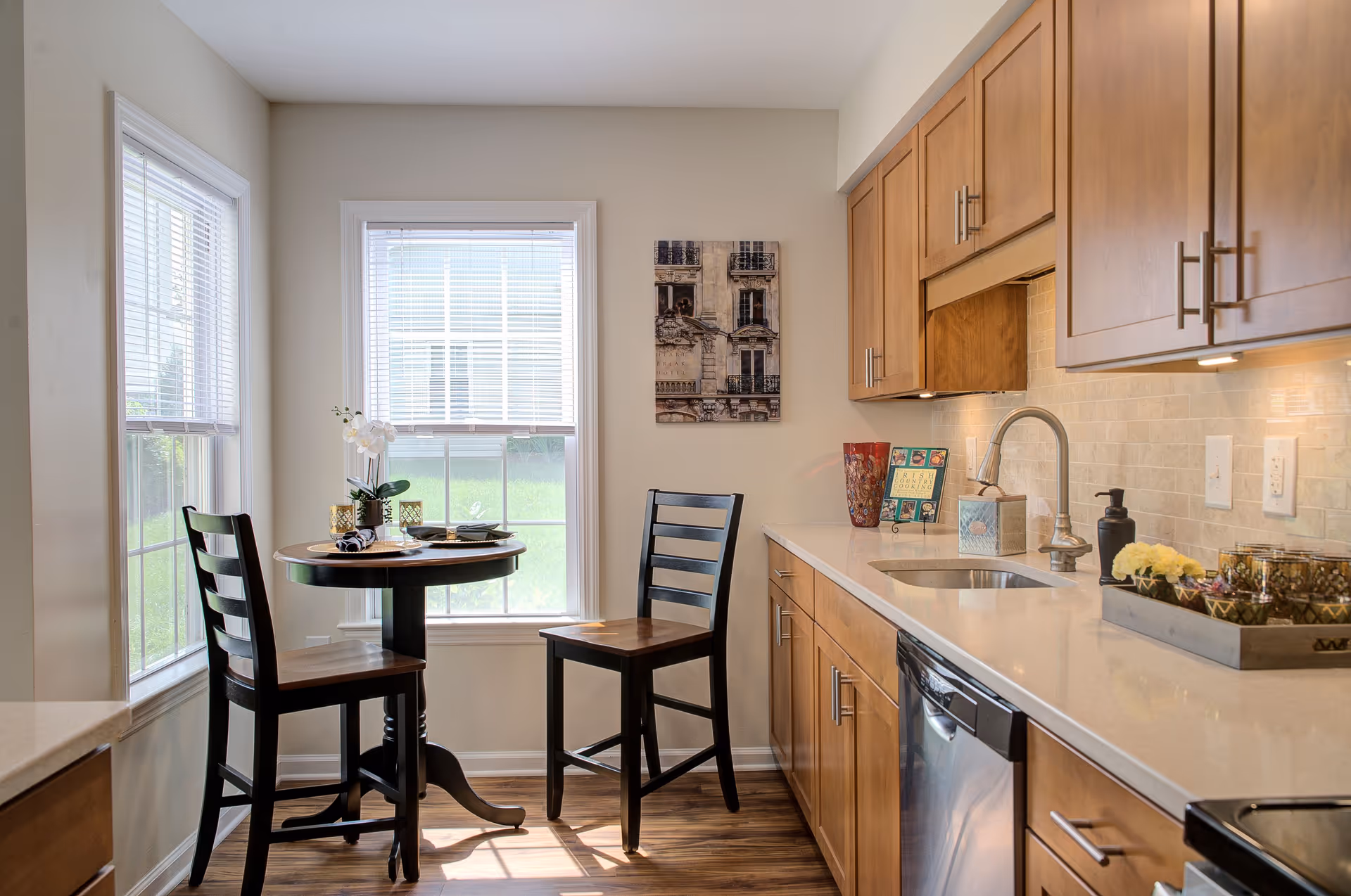 A bright kitchen area with wooden cabinets, a white countertop, and a stainless steel sink and dishwasher. There is a small round wooden table with two chairs near two large windows with white blinds. The table is set with plates, napkins, and a small potted orchid. A framed picture hangs on the wall above the table.