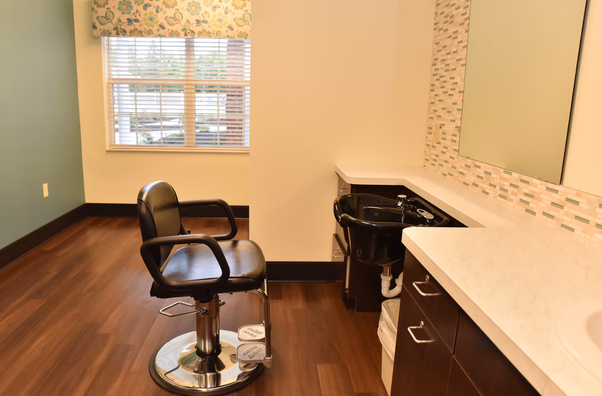 A hair salon station inside a senior living facility with a black salon chair, a black wash basin, a large mirror, and a countertop with drawers. The room has wood flooring, a window with floral curtains, and light-colored walls.