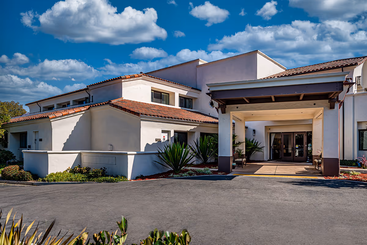 Front entrance of a Mediterranean-style senior living building with a covered porte-cochere, driveway and landscaping under a blue sky.