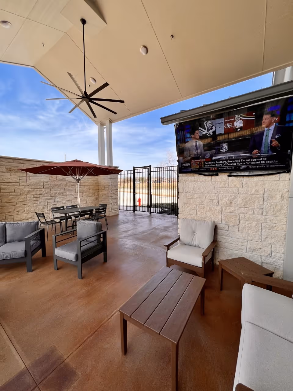 Covered outdoor lounge area with sofas, chairs, a dining table and umbrella, a large ceiling fan, and a wall-mounted TV showing sports.