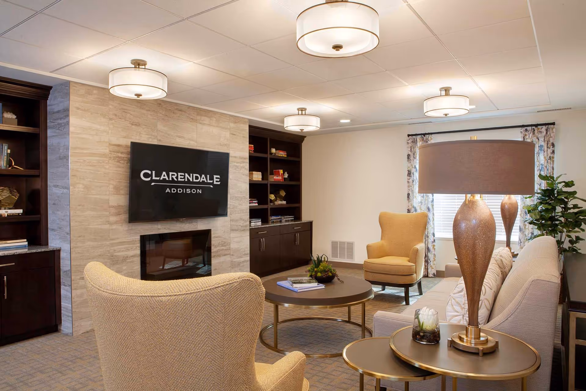A cozy living room area in Clarendale of Addison featuring beige upholstered chairs and sofa, a round coffee table with books and a small plant, a modern fireplace with a mounted TV above displaying the Clarendale Addison logo, built-in dark wood shelves with books and decorative items, and three ceiling lights providing warm illumination.