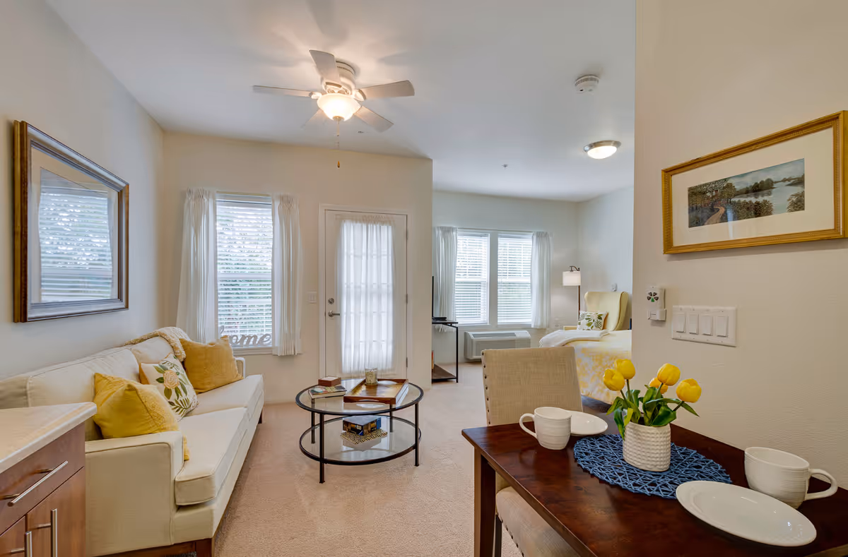Bright studio-style living area with a sofa, round glass coffee table, dining table topped with yellow tulips, and a bed visible in the background.