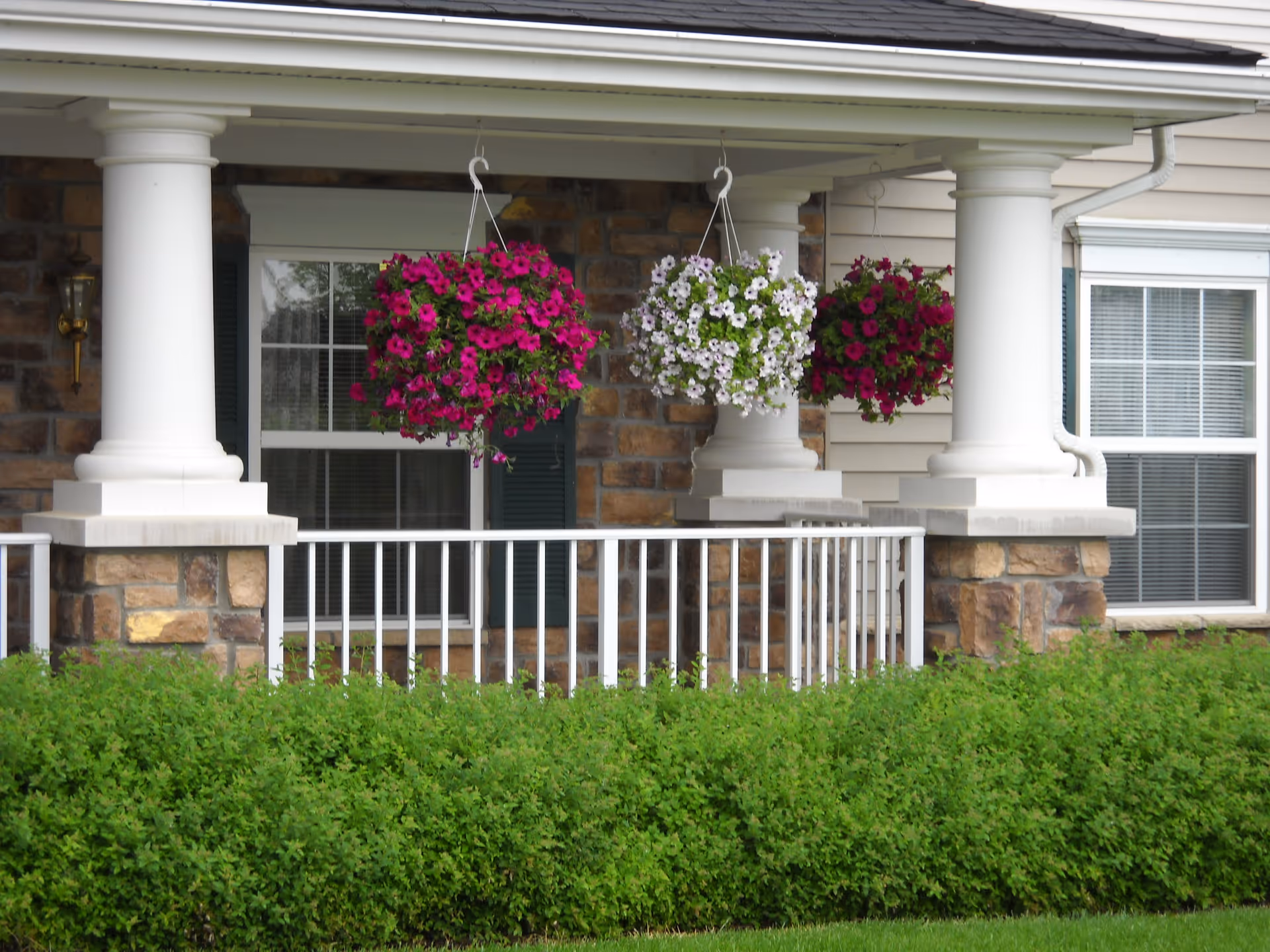 Front porch of a building with white columns and stone accents, featuring three hanging flower baskets with pink and white flowers, white railing, and green shrubs in front.