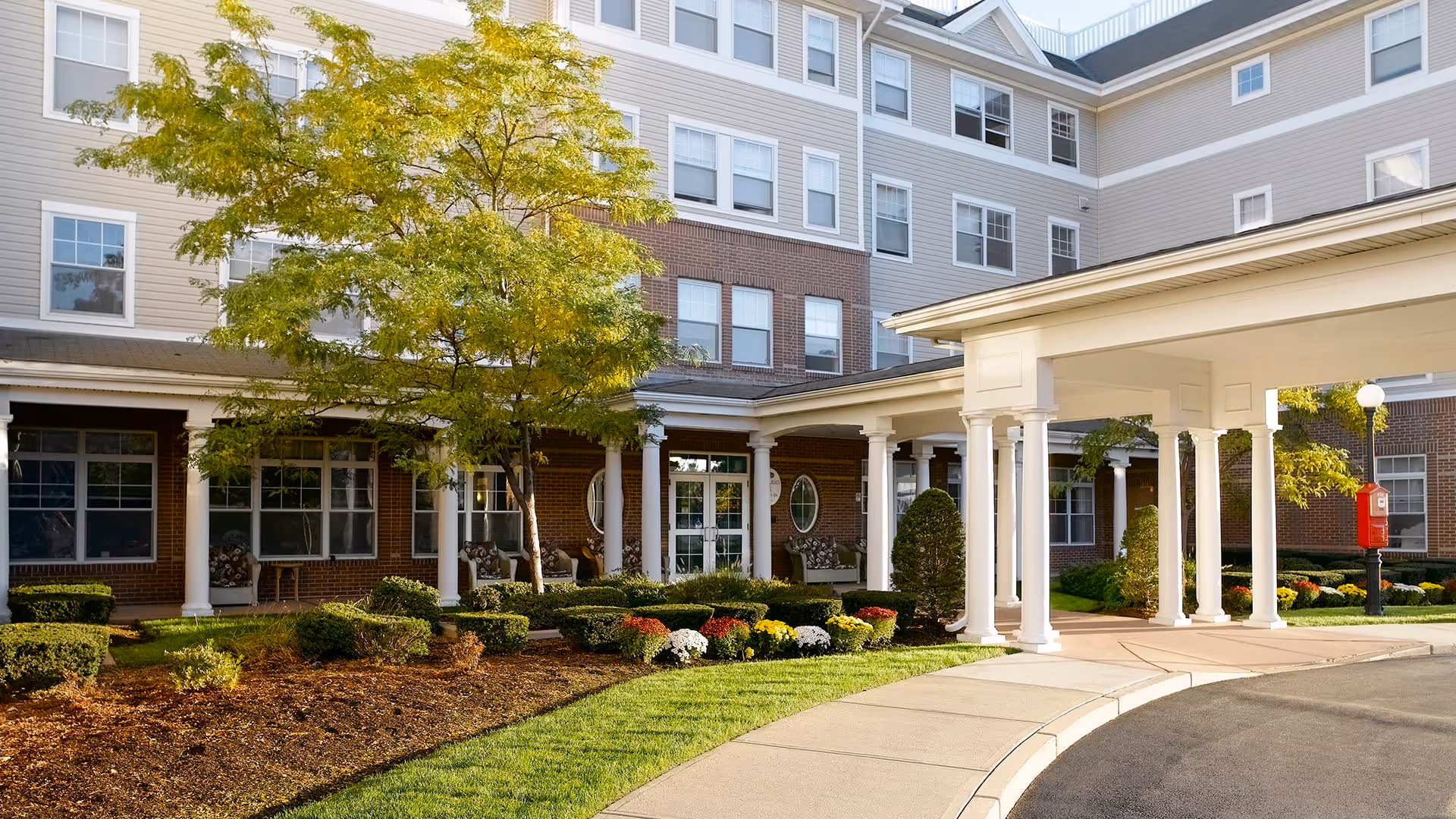 Exterior view of Atria Marina Place senior living facility showing a multi-story building with beige and brick facade, white columns supporting a covered entrance, landscaped garden with trimmed bushes, flowers, and a tree, and a curved driveway.