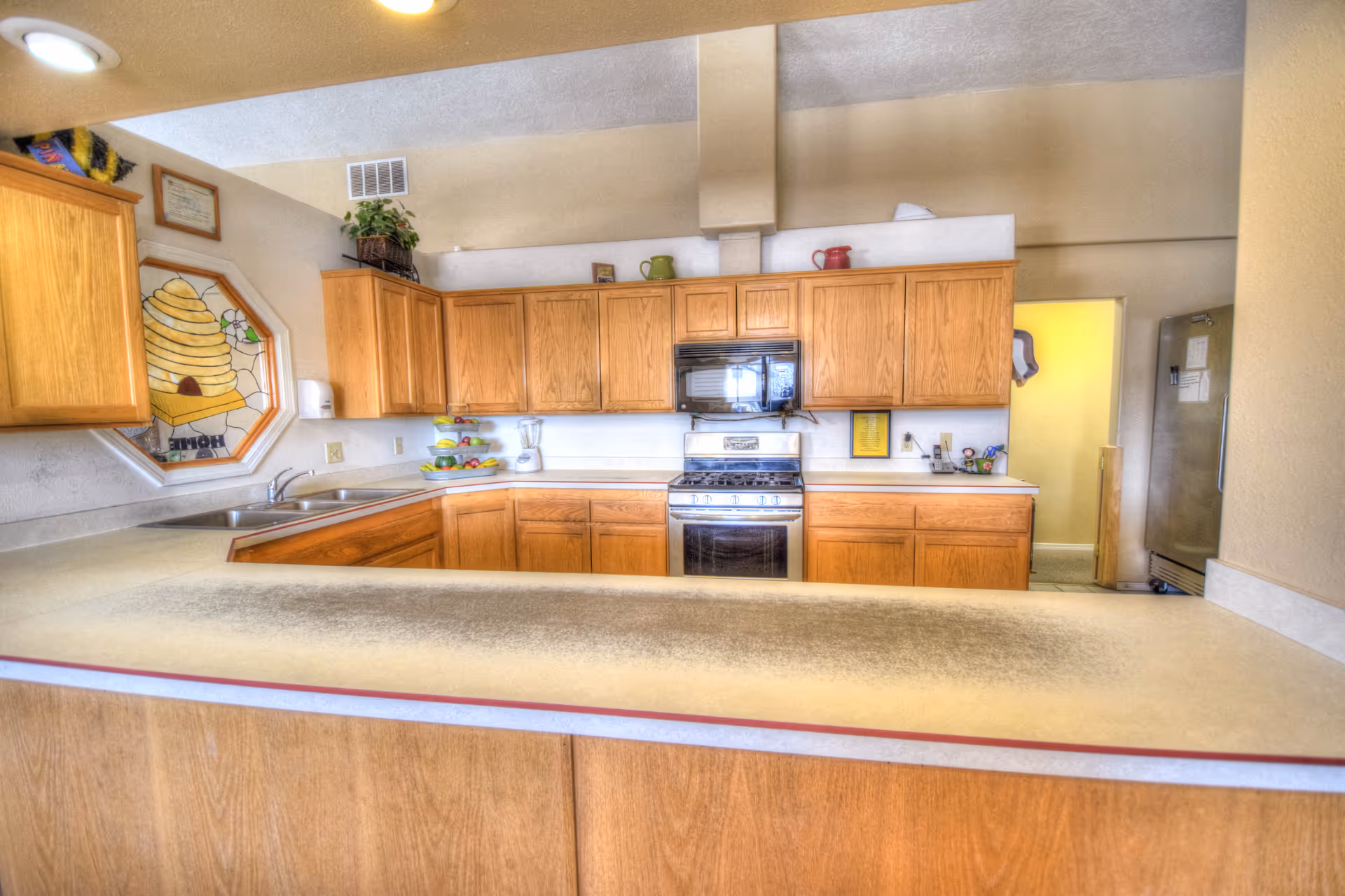 A kitchen with wooden cabinets, a stove with oven, a microwave above it, a double sink, a refrigerator, and a countertop in the foreground. There is a decorative stained glass window with a beehive design on the left wall and some kitchen items like a blender and fruit bowl on the counters.