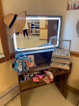 A wooden vanity table with a large mirror surrounded by LED lights. On the table are colorful scarves and an open jewelry box with various necklaces and bracelets. A straw hat hangs on the left side of the mirror. The background shows a hallway and part of another room.