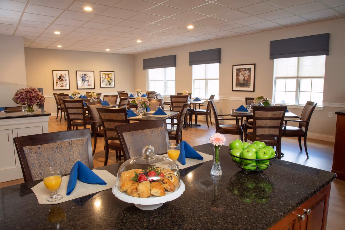 Bright communal dining room with tables set with blue napkins, a counter displaying pastries under a glass dome and a bowl of green apples.