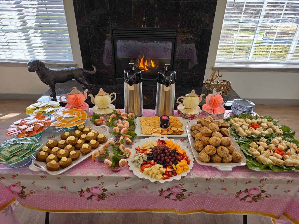 A table set with a variety of snacks and refreshments in front of a fireplace. The table has a floral and pink tablecloth and holds plates of cookies, crackers with cheese, shrimp cocktail, sandwiches, and muffins. There are two coffee dispensers, decorative teapots, and stacks of tea bags. The background shows windows with blinds and a black dog statue on the left side.