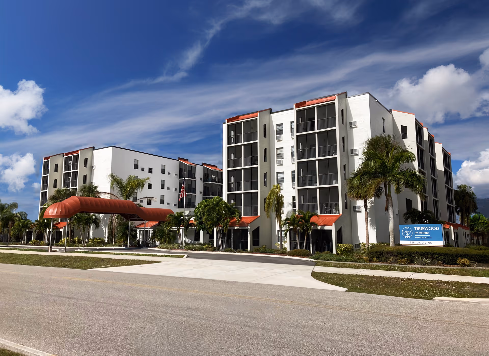 Front exterior of a multi-story senior living building with balconies, red awnings and palm trees under a blue sky.