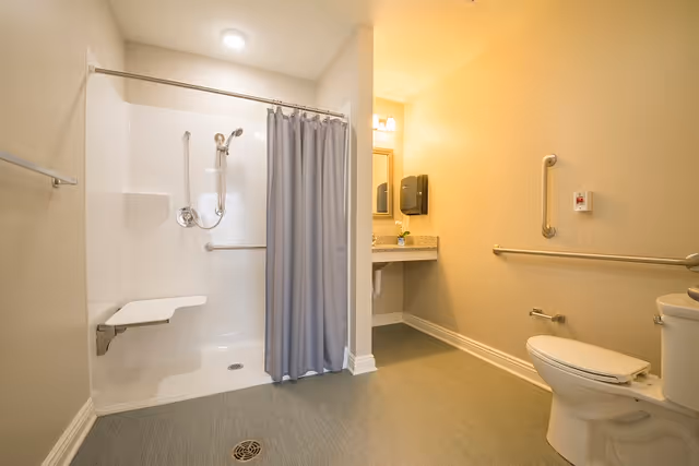 A spacious accessible bathroom with a walk-in shower featuring a fold-down seat and grab bars, a toilet with grab bars on the adjacent walls, and a vanity with a sink, mirror, and paper towel dispenser. The walls are painted light beige and the floor has a textured surface for safety.