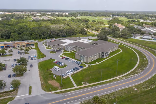 Aerial view of Twin Creeks Assisted Living & Memory Care facility showing a large two-story building with a parking lot, surrounding green lawns, and a curved road in front. The area around the facility includes trees, other buildings, and a distant horizon under a clear sky.