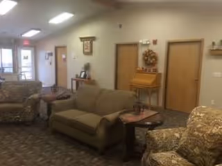 Communal living room with a sofa and patterned armchairs, side tables, and doors with wall decor in the background.