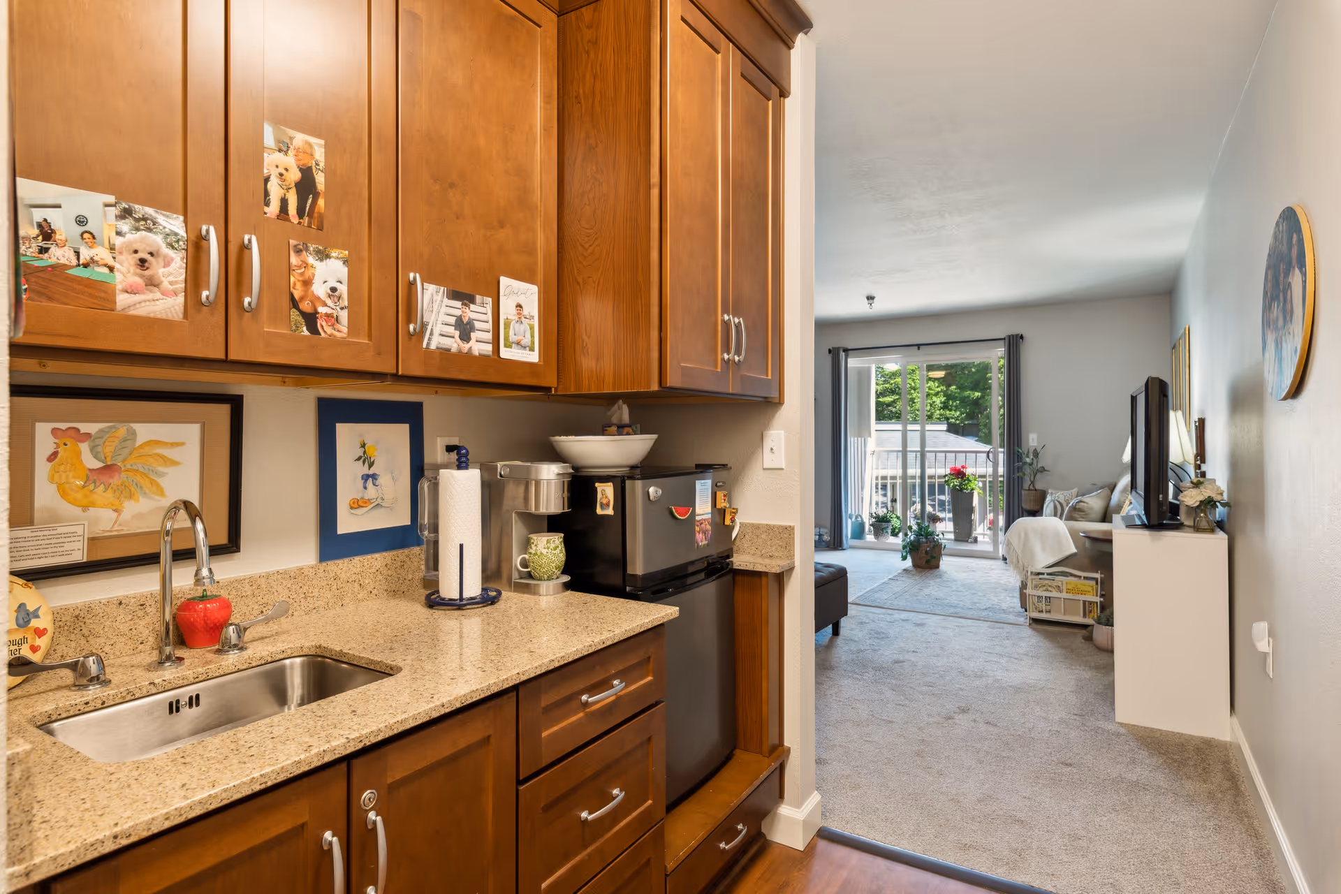 A kitchenette with wooden cabinets, a sink, and countertop appliances opening into a carpeted living room with a sliding glass door to a balcony.