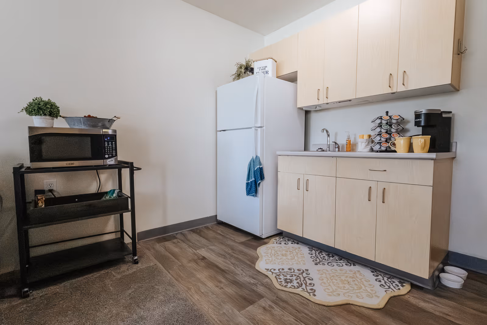 A small kitchen area with light wood cabinets, a white refrigerator, a sink, and a coffee maker with mugs on the countertop. A microwave sits on a black metal cart with shelves, and there is a decorative rug on the floor in front of the cabinets.