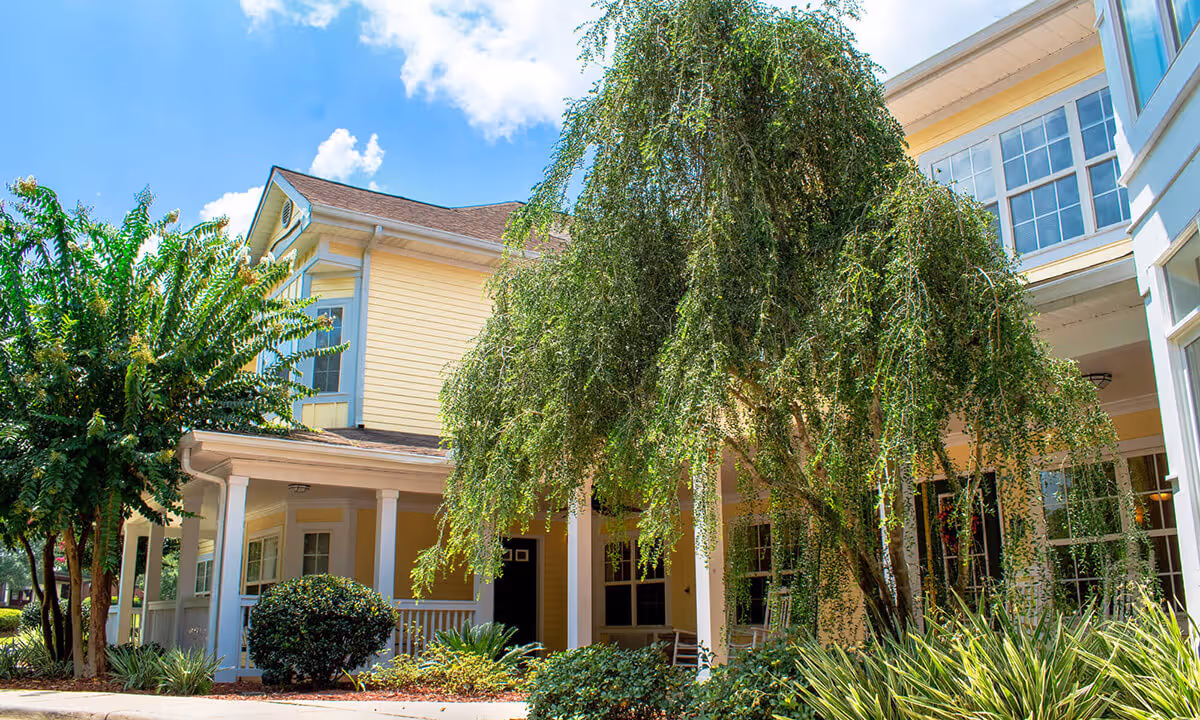 Yellow two-story building front with a covered porch, lush trees and landscaped grounds under a blue sky.