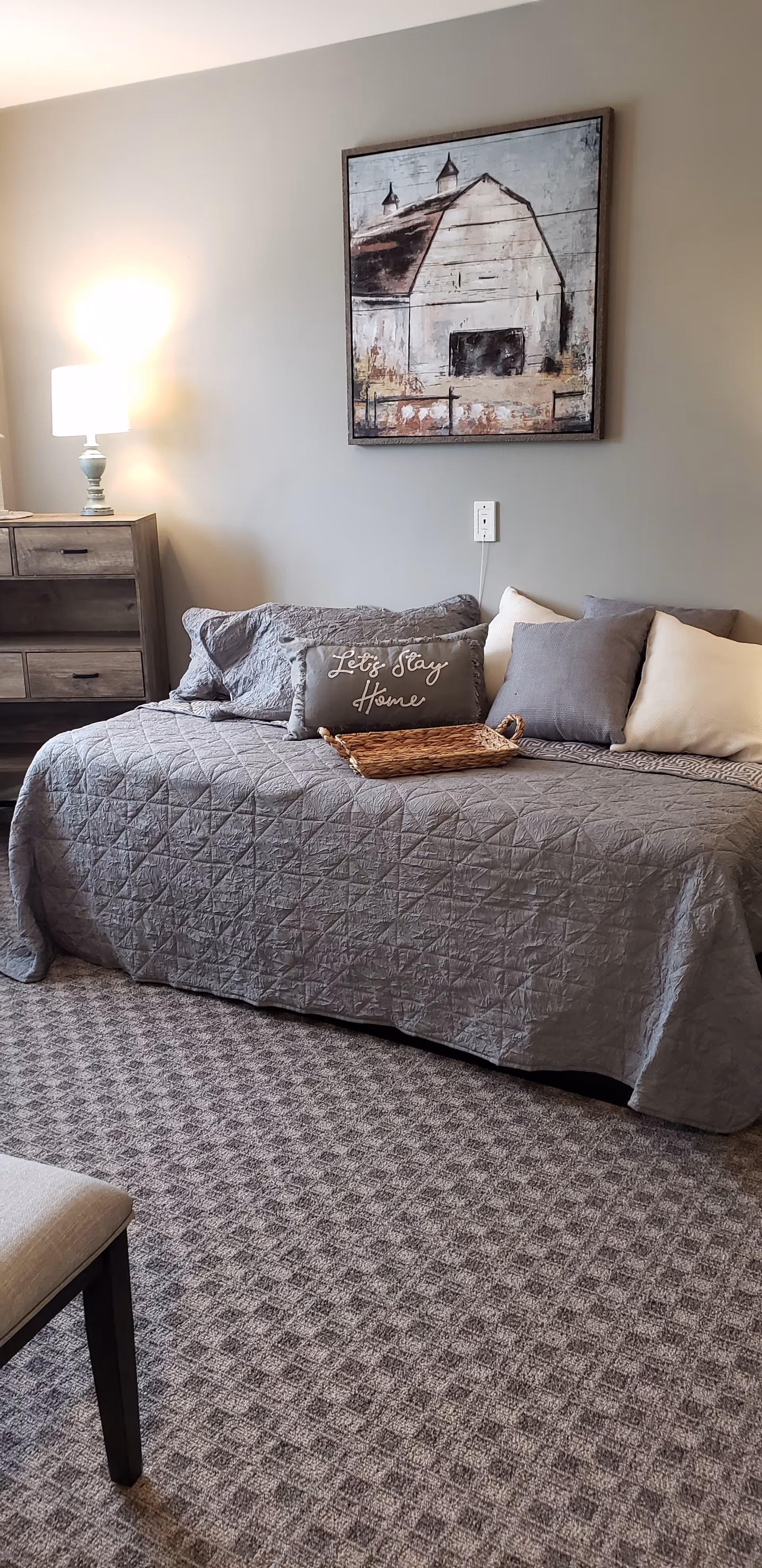 A cozy bedroom with a gray quilted bedspread on a bed, several pillows including one with the text 'Let's Stay Home', a wicker tray on the bed, a wooden dresser with a lamp on top, and a framed painting of a barn on the wall above the bed.