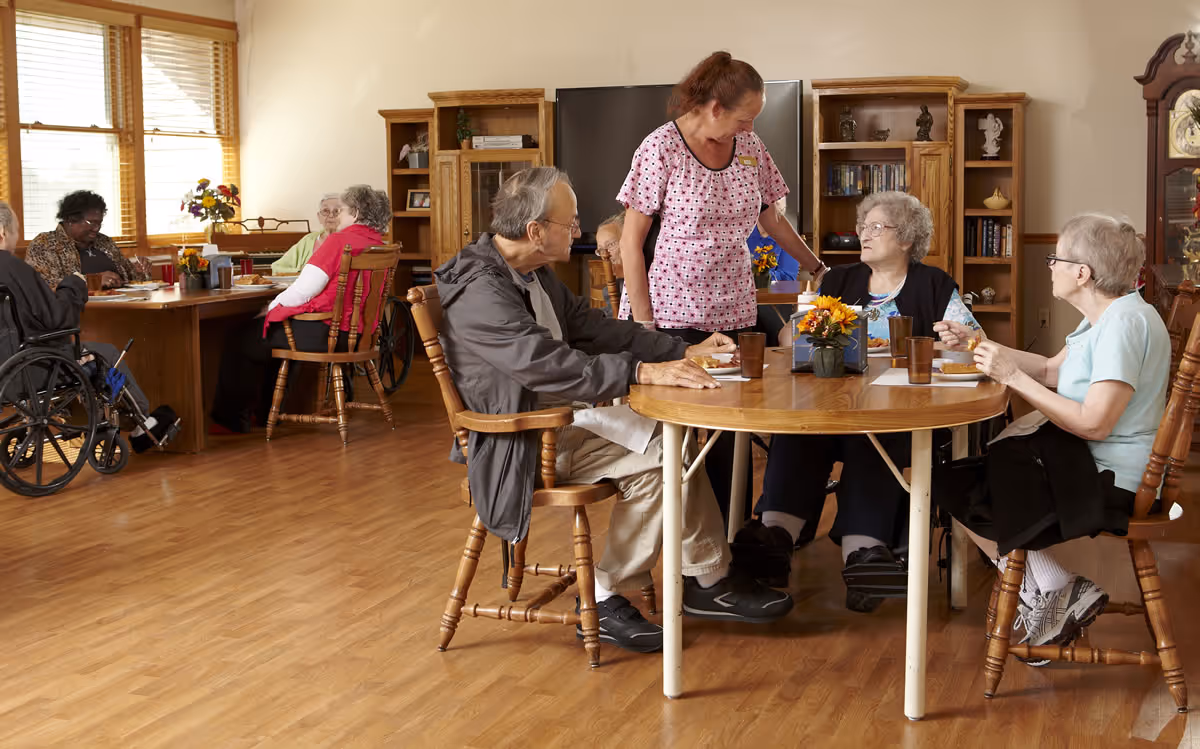Elderly residents and a caregiver seated around tables in a communal dining room of an assisted living facility.