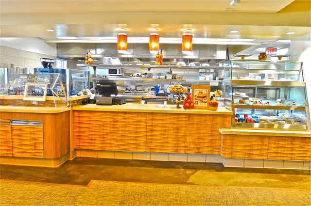 A bright and clean cafeteria counter with wooden paneling, glass display cases filled with pastries and desserts, a coffee sign, and kitchen equipment visible in the background under warm hanging lights.