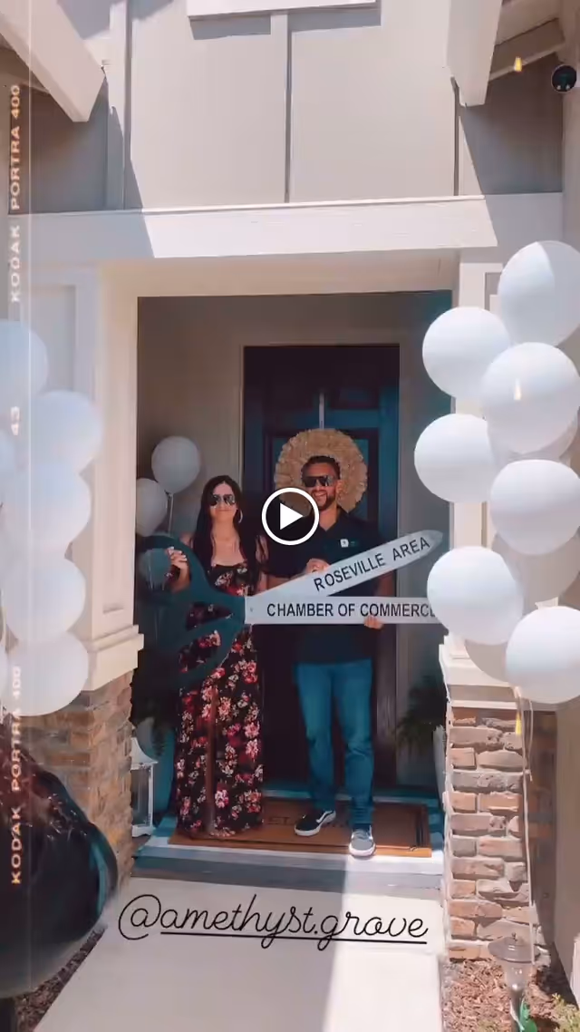 Two people standing at the entrance of a building decorated with white balloons. They are holding a large ceremonial pair of scissors and a sign that reads 'Roseville Area Chamber of Commerce'. The entrance has stone pillars and a dark front door with a wreath.