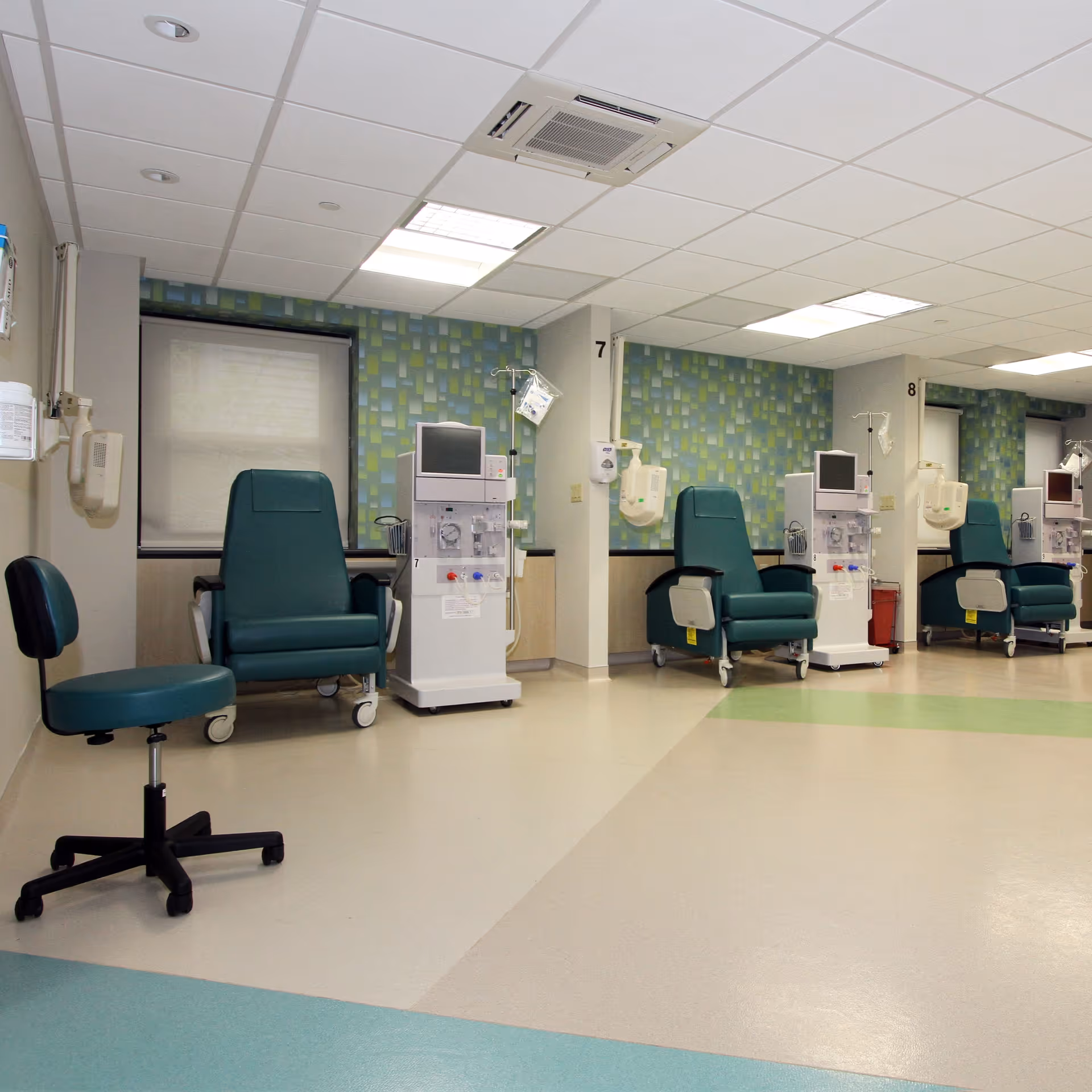 A medical treatment room with multiple green reclining chairs and dialysis machines lined up against a wall with patterned wallpaper. The room has a clean, clinical appearance with a tiled ceiling and bright lighting.