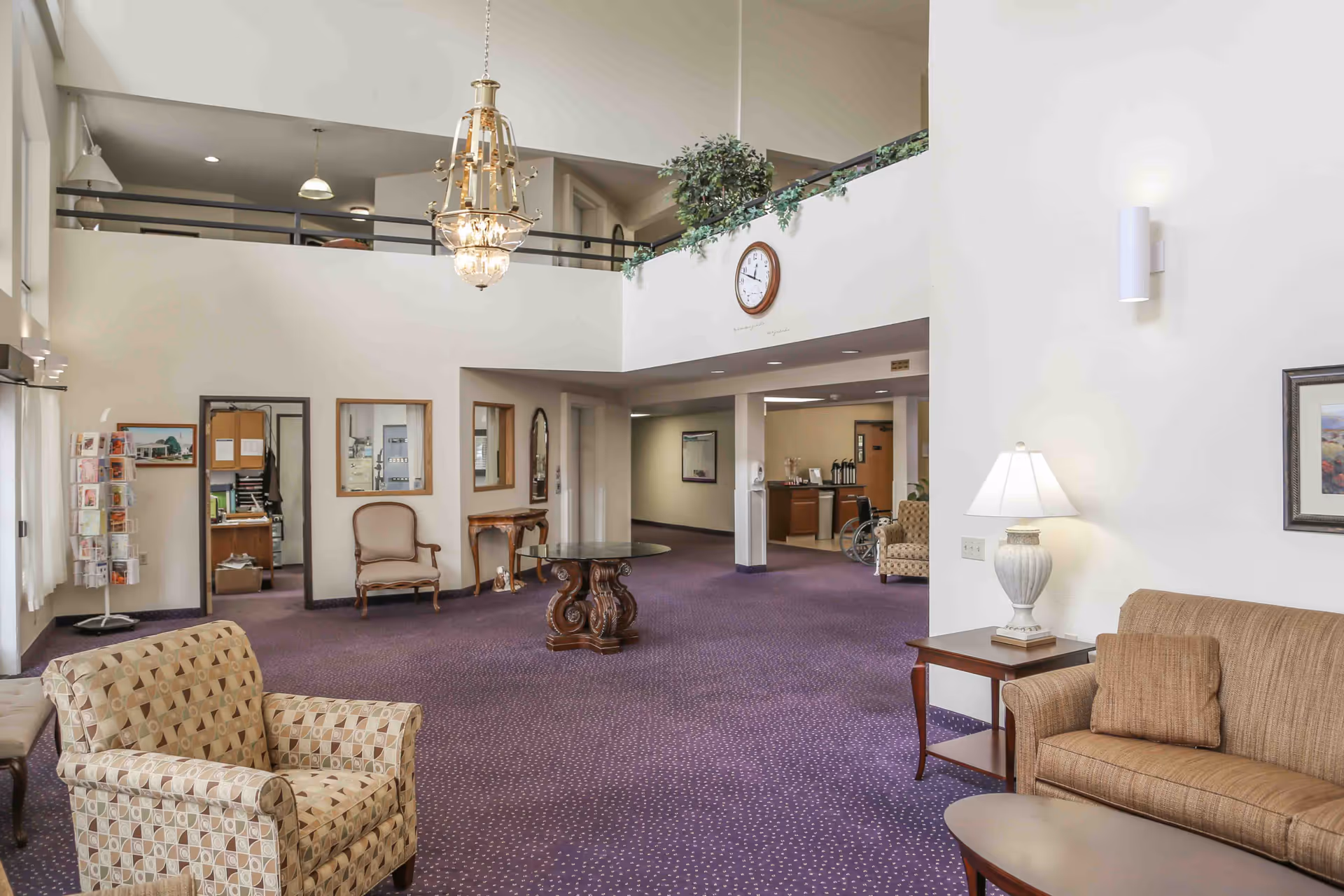 Spacious two-story senior living lobby with upholstered chairs, a central table, chandelier, and a reception area.