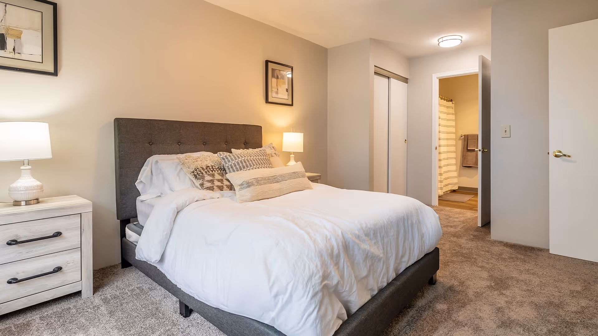 A cozy bedroom with a gray upholstered bed frame, white bedding, and multiple decorative pillows. There are two bedside tables with lamps on each side of the bed. The room has beige walls and carpeted flooring. A doorway leads to a bathroom with a shower curtain and towels visible.