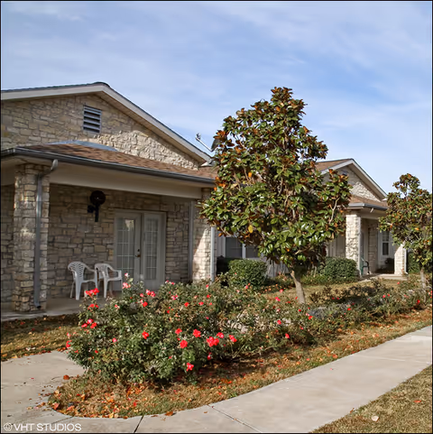 Exterior view of a stone building with a covered porch area featuring white plastic chairs. There are small trees and bushes with red flowers planted along a sidewalk in front of the building under a partly cloudy sky.