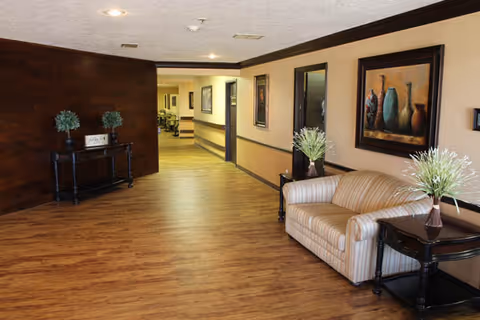 Interior hallway of a nursing and rehabilitation facility with wooden flooring, beige walls, and dark wood trim. A striped beige couch is positioned against the right wall next to a dark wooden side table with a vase of flowers. On the left side, there is a small black console table with two potted plants and a decorative sign. Several framed artworks hang on the walls, and the hallway extends into the distance with more seating visible.