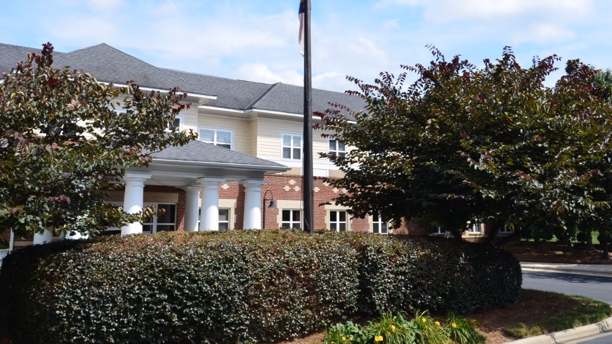 Exterior view of a senior living facility building with brick and light-colored siding, white columns at the entrance, surrounded by well-maintained bushes and trees under a partly cloudy sky.