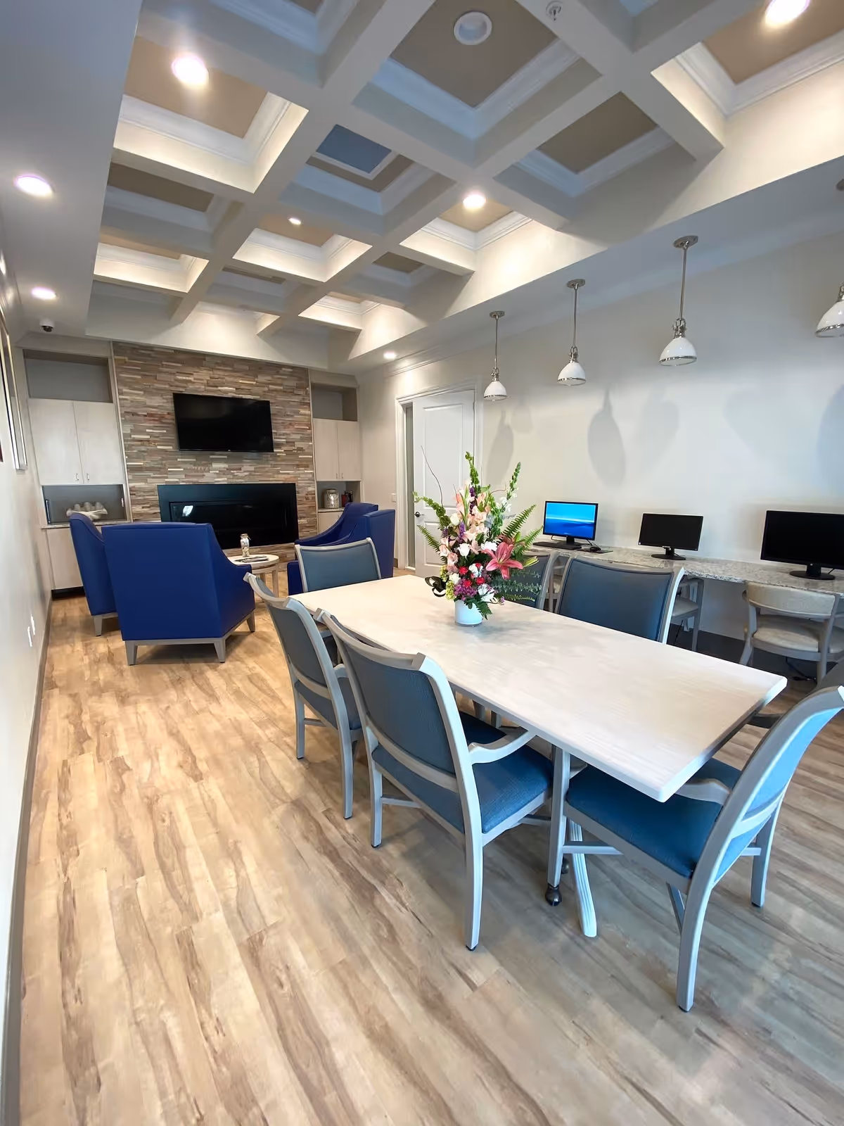 A modern common area with a long table and six chairs, a vase with flowers on the table, three computer stations with monitors along the wall, and a seating area with blue armchairs in front of a stone accent wall with a mounted TV and fireplace. The ceiling features a coffered design with recessed lighting.