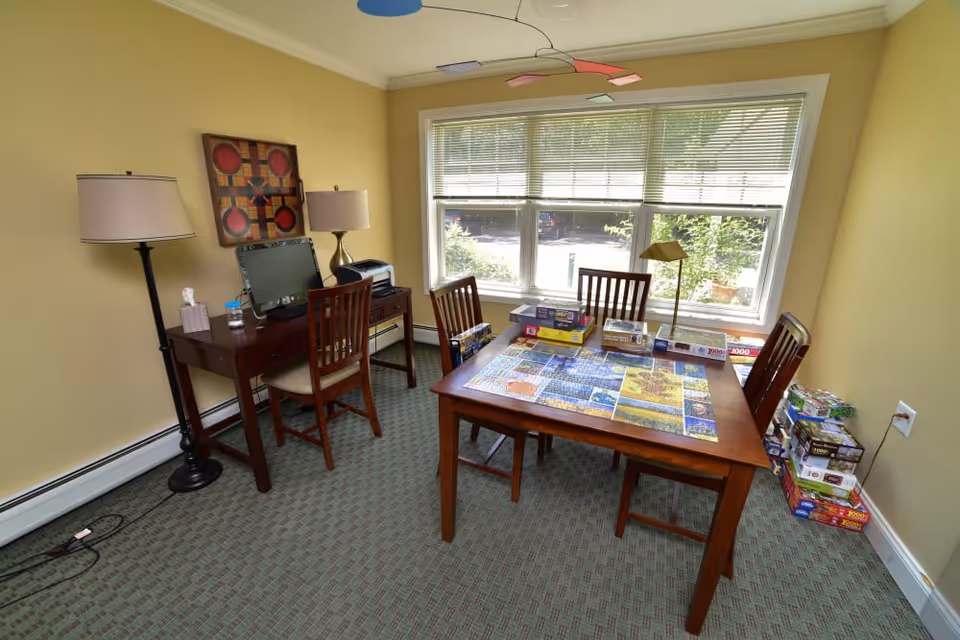 Bright interior activity room with a wooden table mid-way through a jigsaw puzzle, chairs, a desk with a computer and lamps by a large window.