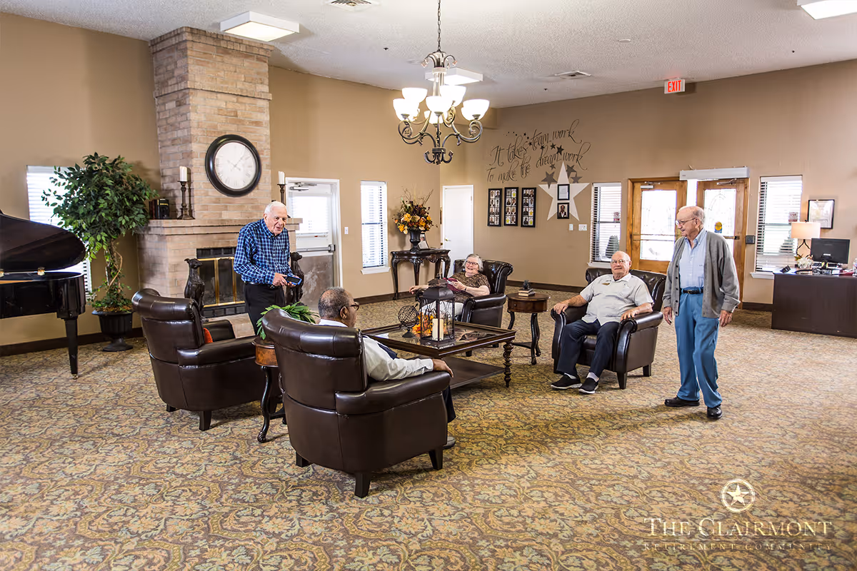 A group of elderly people sitting and standing in a spacious, well-lit common area with beige walls and patterned carpet. The room features a brick fireplace with a clock above it, a grand piano, leather armchairs arranged around a coffee table, and a chandelier hanging from the ceiling. There are windows and a door in the background, along with framed pictures and decorative wall art.