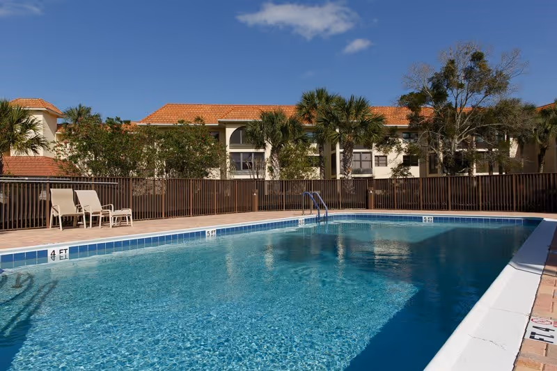 Outdoor swimming pool with clear blue water surrounded by a tiled deck. There are a few lounge chairs on the left side near a brown fence. In the background, there are palm trees and a building with a red-tiled roof under a clear blue sky.