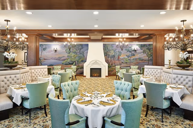 Elegant dining room with round tables covered in white tablecloths, set with plates, napkins, and silverware. The room features tufted light green chairs and beige upholstered booth seating. A white fireplace is centered on the far wall, flanked by a mural depicting a scenic landscape with trees and a house. Ornate chandeliers hang from the ceiling, and the carpet has a floral pattern.