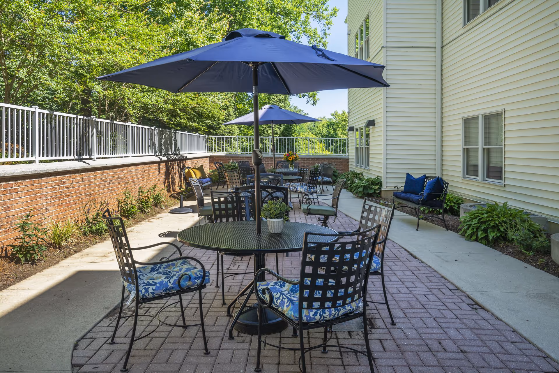 Outdoor patio area with several black metal tables and chairs with blue patterned cushions. Each table has a large blue umbrella providing shade. The patio is paved with bricks and surrounded by a low brick wall and greenery. The side of a light yellow building with windows and some plants along the walkway is visible.
