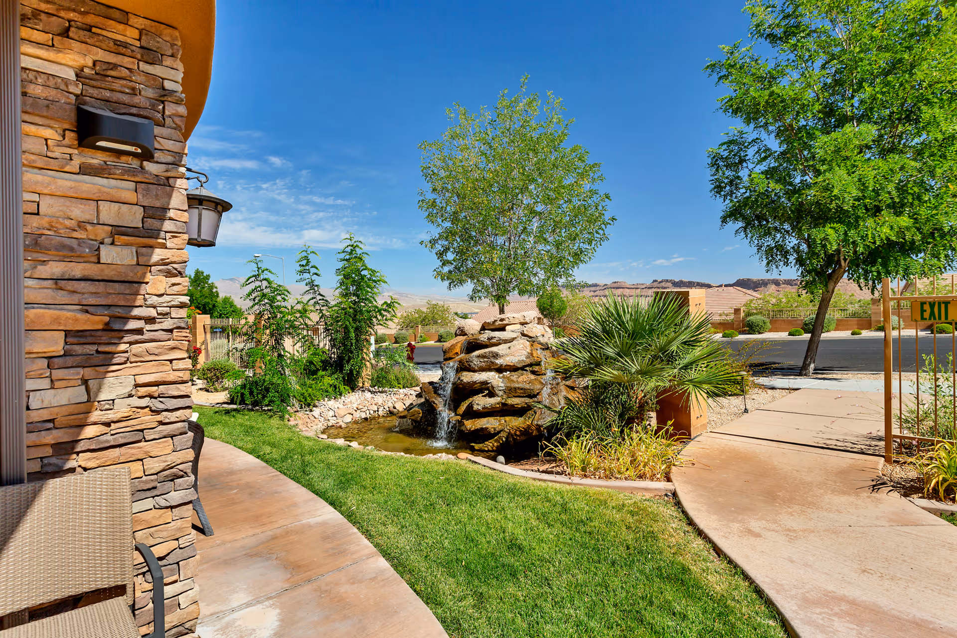 Outdoor area at The Retreat at Sunriver featuring a small waterfall flowing over rocks into a pond, surrounded by green grass, plants, and trees with a clear blue sky in the background. A stone wall and patio with chairs are visible on the left side, and a paved walkway leads to an exit gate on the right.