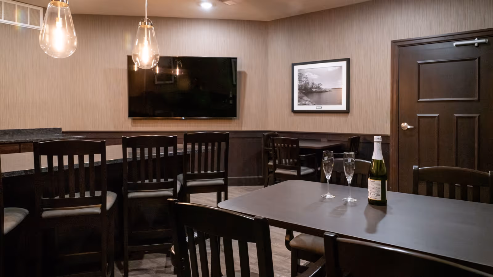 A dining area with dark wooden chairs and tables. On one table, there is a bottle of champagne and two filled champagne glasses. The room has beige walls with dark wood paneling, a flat-screen TV mounted on the wall, a framed black and white photograph, and two hanging pendant lights.