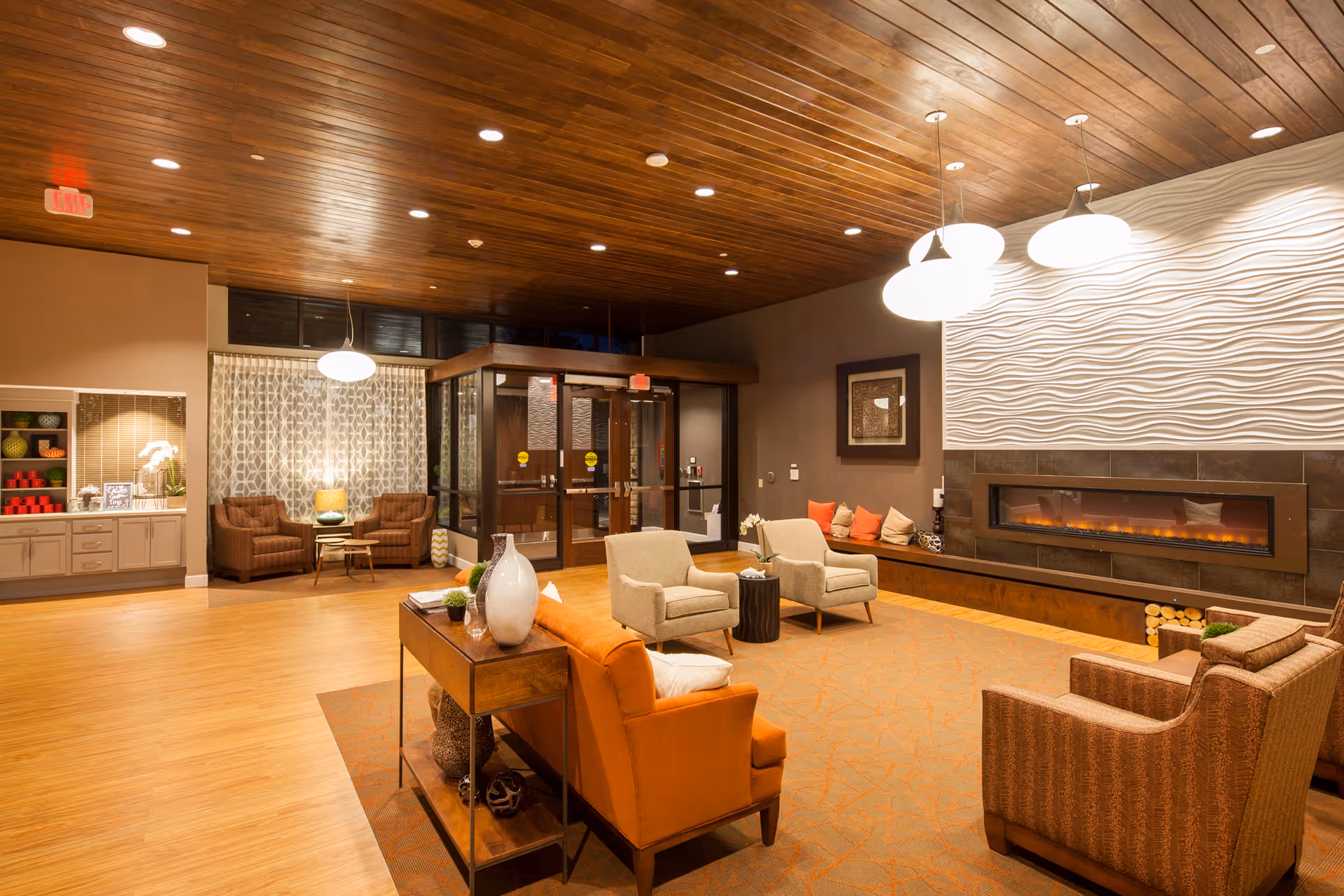 A cozy and well-lit living room area in a senior living facility featuring a modern electric fireplace with a textured white wall above it. The room has a wooden ceiling with recessed lighting, a wooden floor, and comfortable seating including an orange sofa, beige armchairs, and patterned chairs. There is a small table with decorative items and a cabinet with colorful decor on the left side. Glass doors lead to another area in the background.