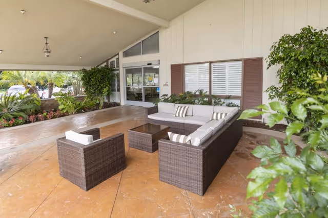 Outdoor covered seating area with a wicker sectional sofa, armchair, and glass-top coffee table on a tiled floor near the entrance of a building surrounded by plants and greenery.